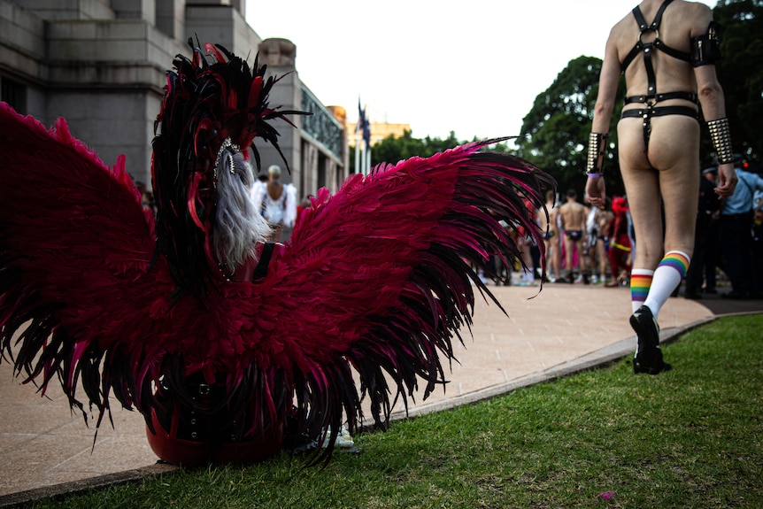 Somebody sits in a feather-covered costume as a man, dressed in leatherware with buttocks exposed, walks away
