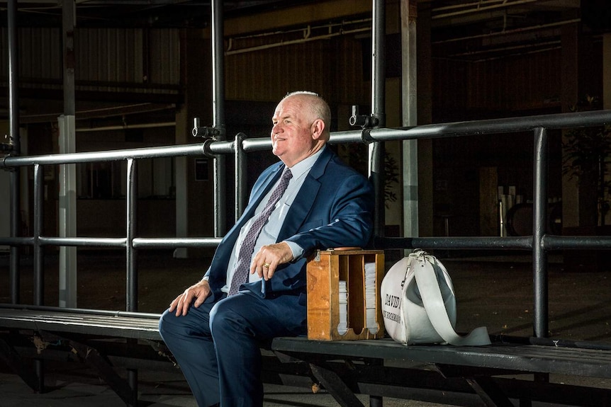 David Dwyer sits on a bench with his bag next to him, looking into the distance.