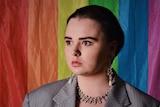 Kirra Hampson wears a grey pinstripe suit, earrings and a necklace and stands against a rainbow flag.