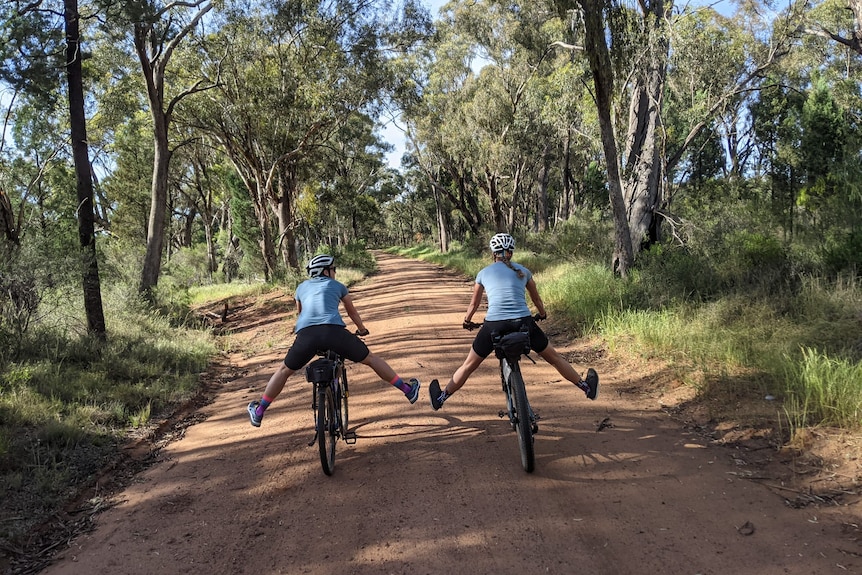 Two women stick their legs out while on bikes on a fire trail.