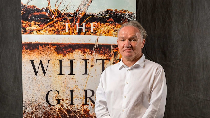 An Aboriginal man in his late 50s in a white shirt stands in front of a poster with the words 'The White Girl'