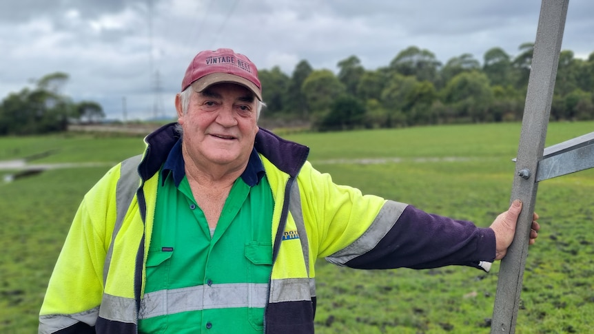 Old man dressed in high-vis and a cap leans on a power pole in an emerald green paddock