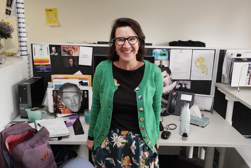 A smiling white woman in a black top and green cardigan stands in front of a desk decorated with photos of Benedict Cumberbatch.