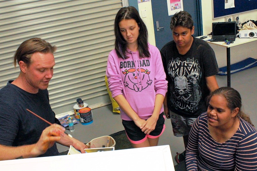 Three young people watch as the artists shows them how to apply paint