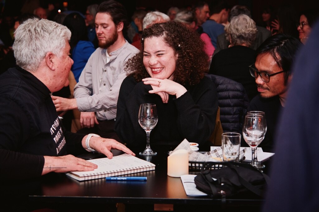 Smiling woman with dark curly hair sitting with a person with grey hair. WIne glasses and a notepad on table.