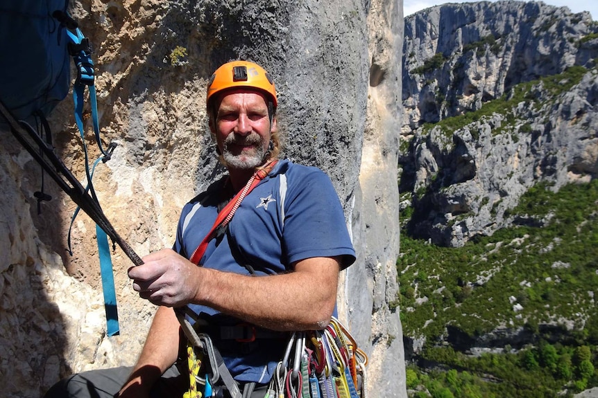 A man in his sixties in rock climbing gear, standing on a ledge on a cliff, looking to camera.