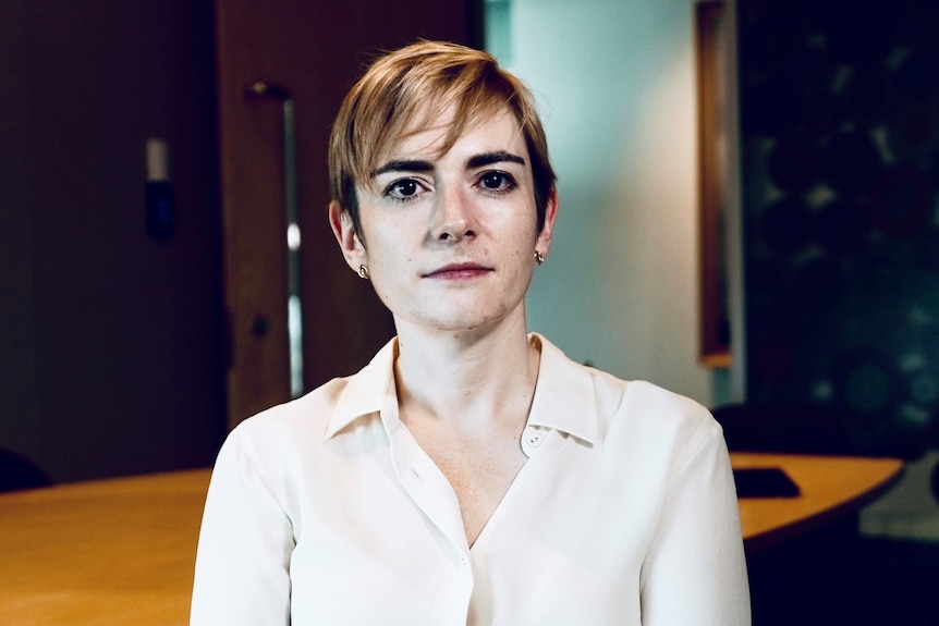 A woman with very short, fair hair, wearing a cream silk shirt looks into the camera.