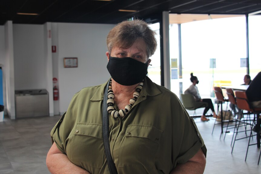 An older woman wearing a facemask smiles at the camera with an airport waiting area behind her
