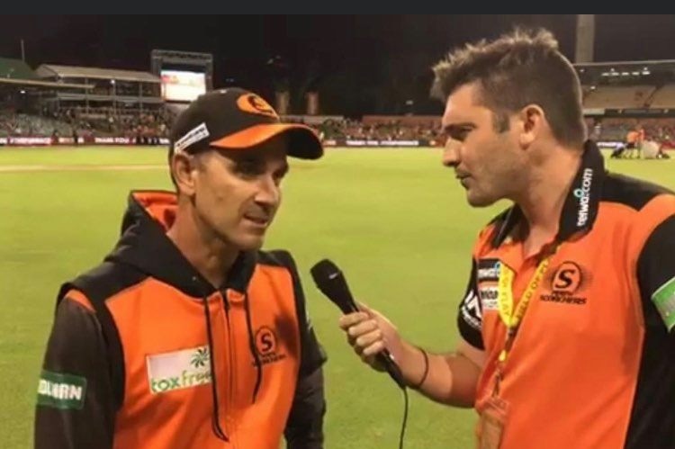 Two men wearing orange stand on a green cricket oval speaking over a microphone