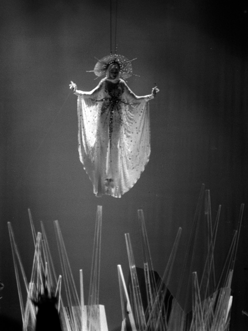A black and white photo of a model dressed as the madonna hanging in the air, pointing see through objects on the ground
