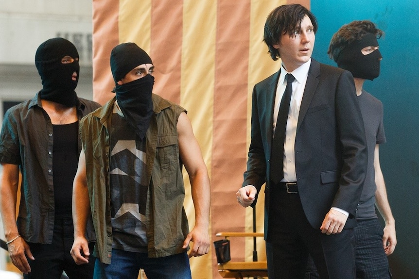 Actor Paul Dano and three masked vigilantes standing up with their heads turned to the right.