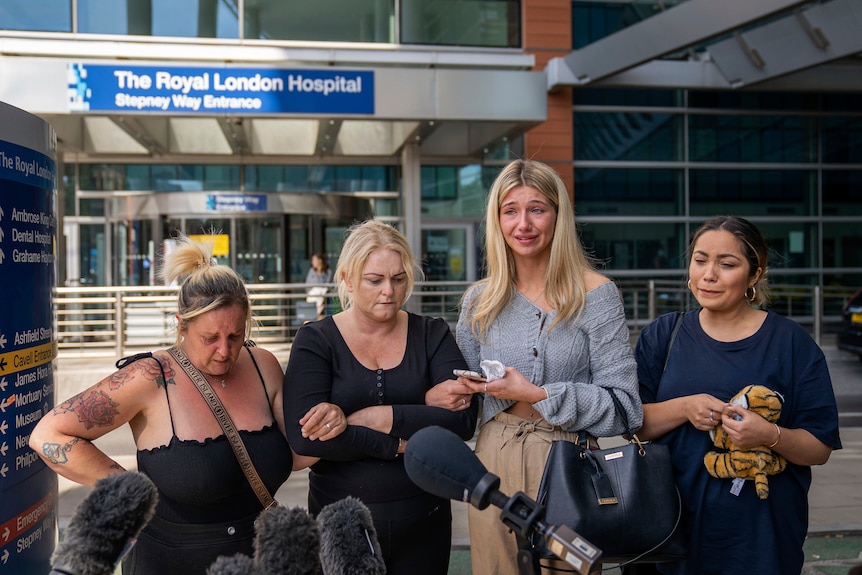 Three women stand with Hollie Dance in front of microphones outside The Royal London Hospital