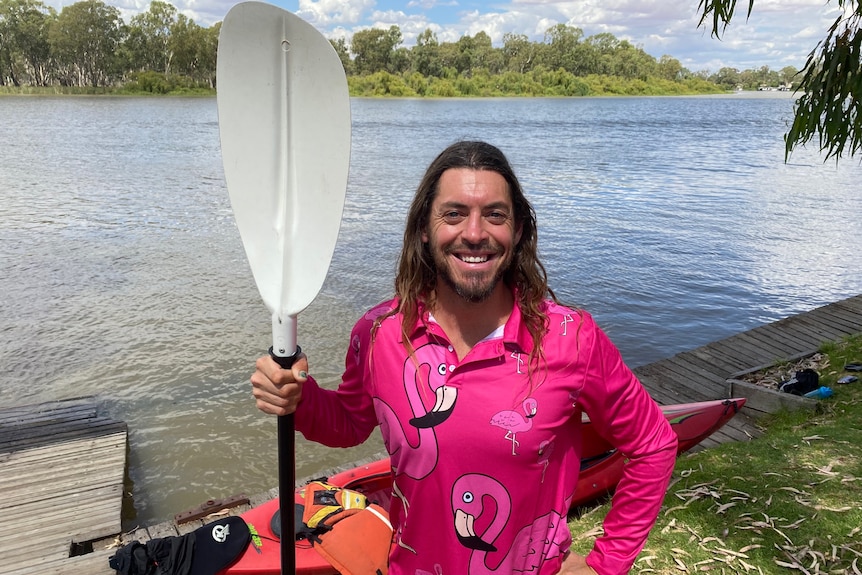 A man wearing a pink shirt holds a kayak paddle in front of a river. 