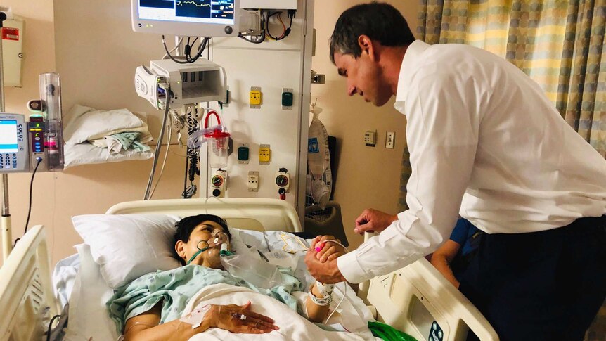 In a hospital room, Beto O'Rourke meets a woman in green hospital garments and holds her hand as she is hooked up to a monitor.