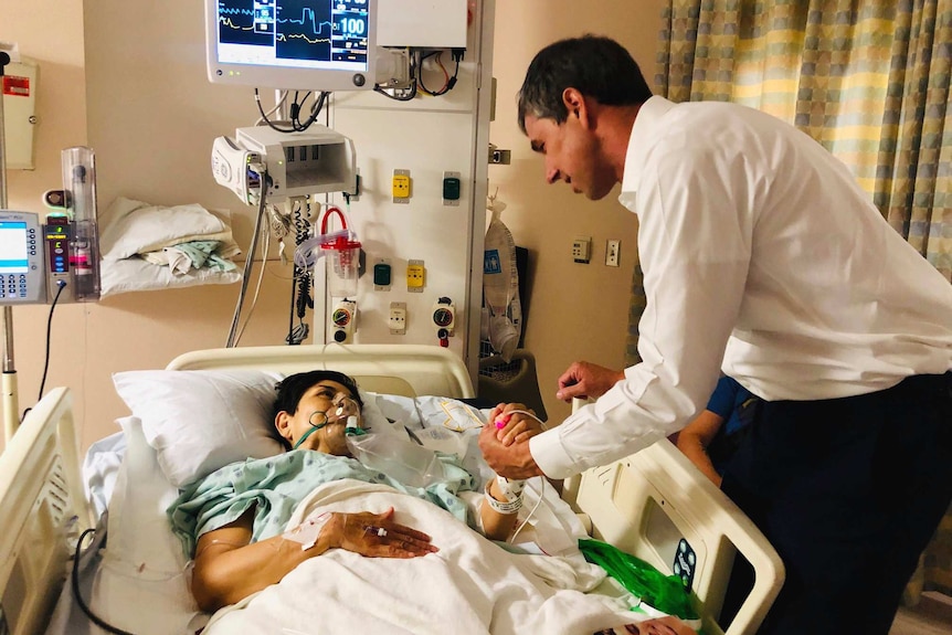 In a hospital room, Beto O'Rourke meets a woman in green hospital garments and holds her hand as she is hooked up to a monitor.