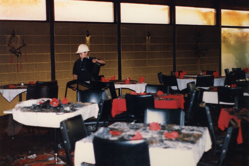 A man with a hard hat moves furniture inside the dining room of a restaurant. Windows, tables and chairs are burnt from a fire