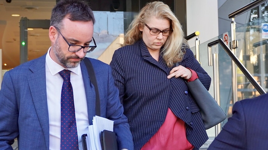 A blonde, bespectacled woman casts her gaze downward as she leaves a court building, flanked by a lawyer in a suit.