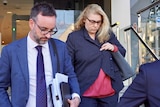 A blonde, bespectacled woman casts her gaze downward as she leaves a court building, flanked by a lawyer in a suit.