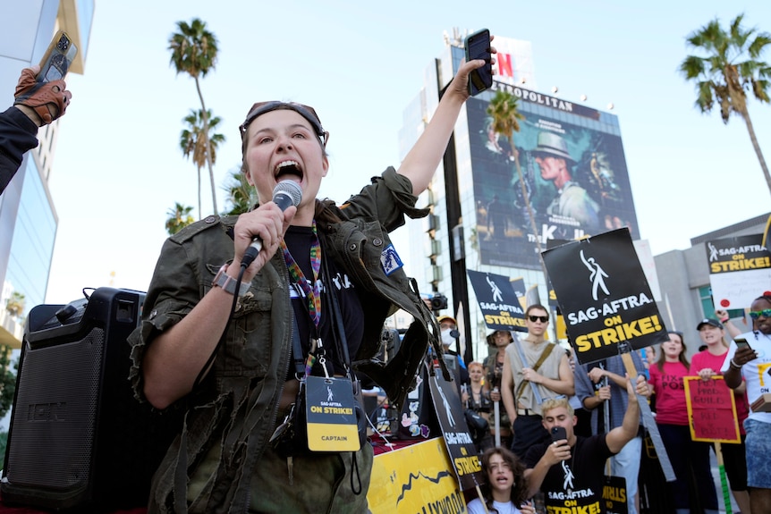 A woman shouts into a microphone in front of an assorted crowd holding SAG-AFTRA on Strike signs
