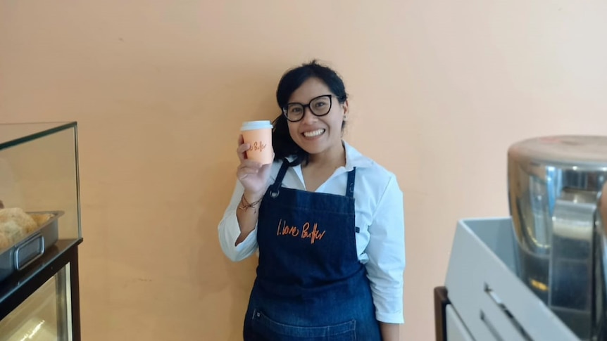 An Indonesian woman smiling while wearing a barista apron and holding a cup of coffee. 