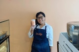 An Indonesian woman smiling while wearing a barista apron and holding a cup of coffee. 
