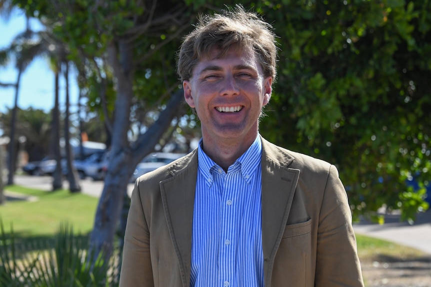 A smiling man wearing a jacket with a shirt underneath stands in front of some trees. 