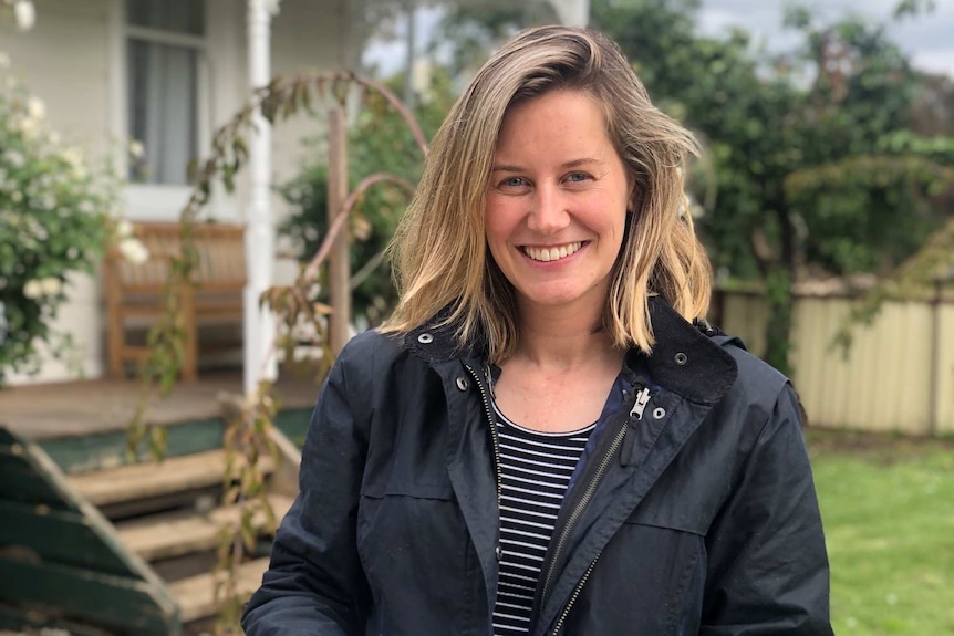 A woman with blonde hair stands in a backyard smiling during the day.