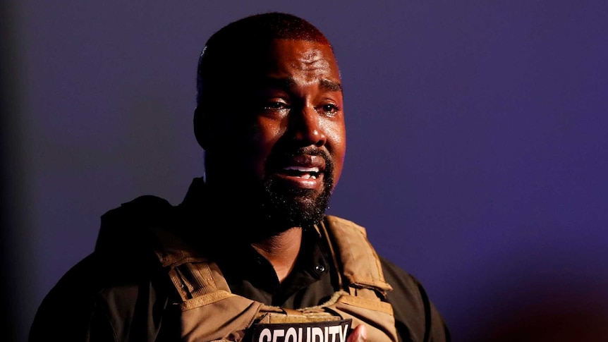 Kanye West holds his hand to his heart with tears in his eyes, wearing a military vest with the word "security" on it