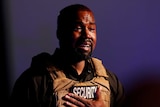 Kanye West holds his hand to his heart with tears in his eyes, wearing a military vest with the word "security" on it