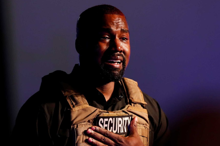Kanye West holds his hand to his heart with tears in his eyes, wearing a military vest with the word "security" on it