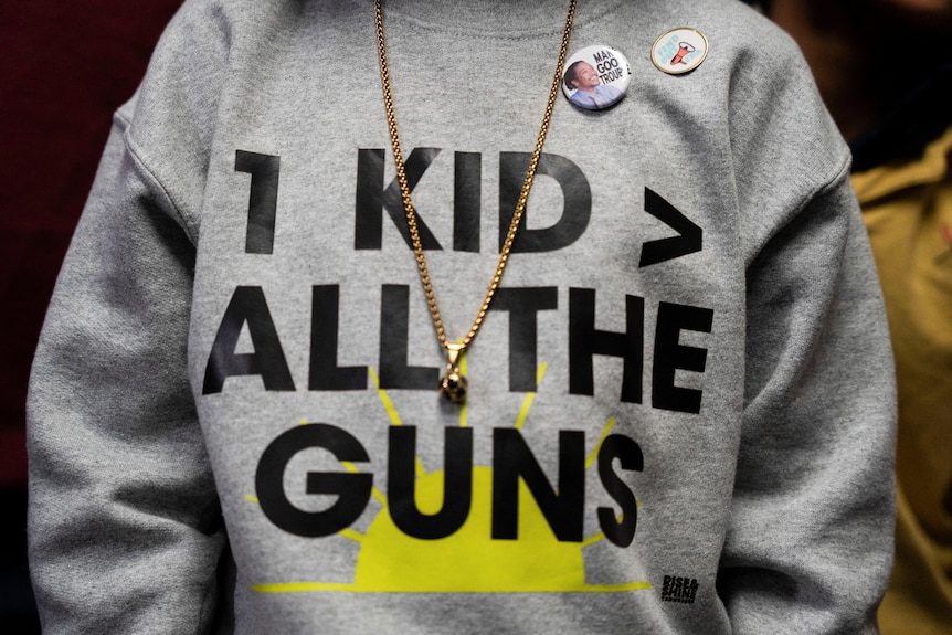 A person wearing a grey sweatshirt with the words 1 Kid greater than all the guns