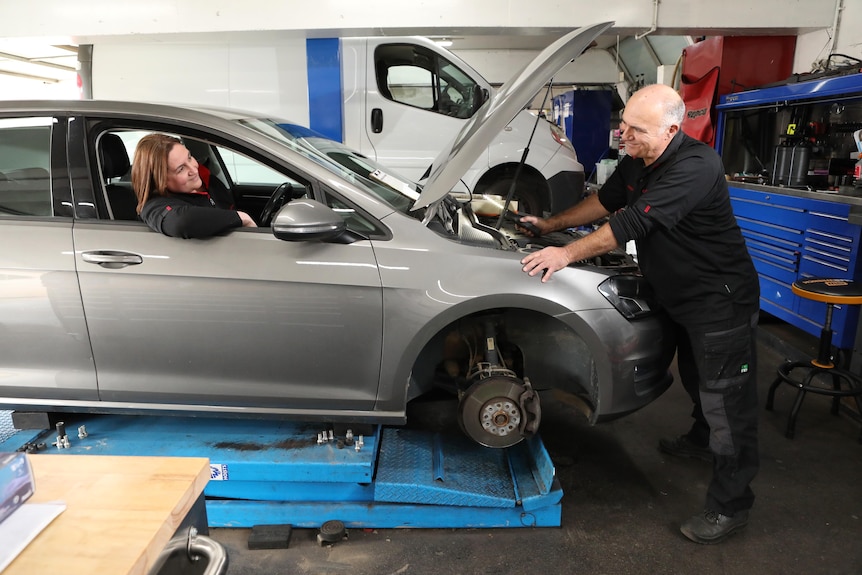 A woman pretends to drive a car at a mechanic while her husband looks under the bonnet.