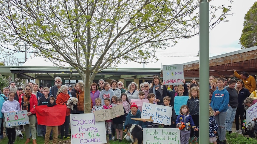 A group of people stand in front of a building holding signs that read social housing not dollars for council