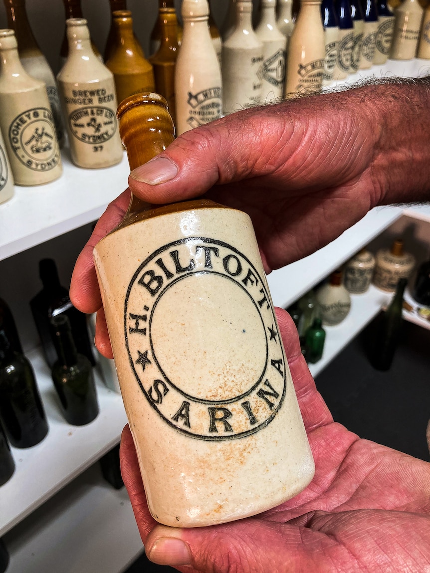 A hand holding a cream ceramic bottle with a brown neck with a shelf stacked full of bottles in the background.