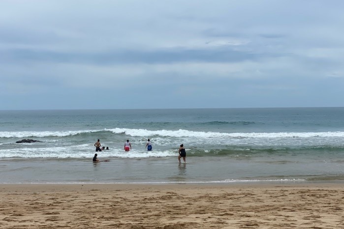 Six people walk through shallow water at the beach into bigger waves.