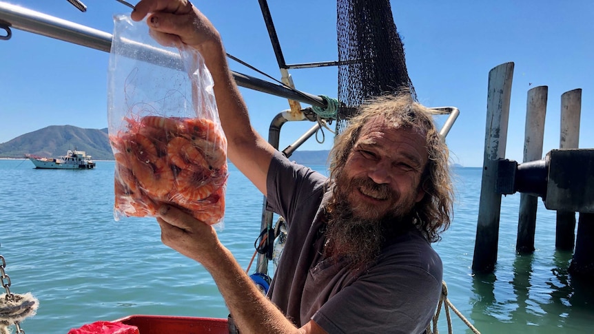 Prawn trawlerman holds up a 1kg bag of freshly caught, cooked tiger prawns with the Cooktown wharf and harbour in the background