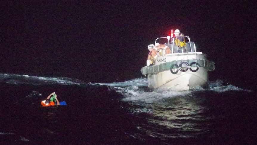 A boat with the Japan Coast Guard pulls alongside a person in a life jacket floating in the ocean at night.