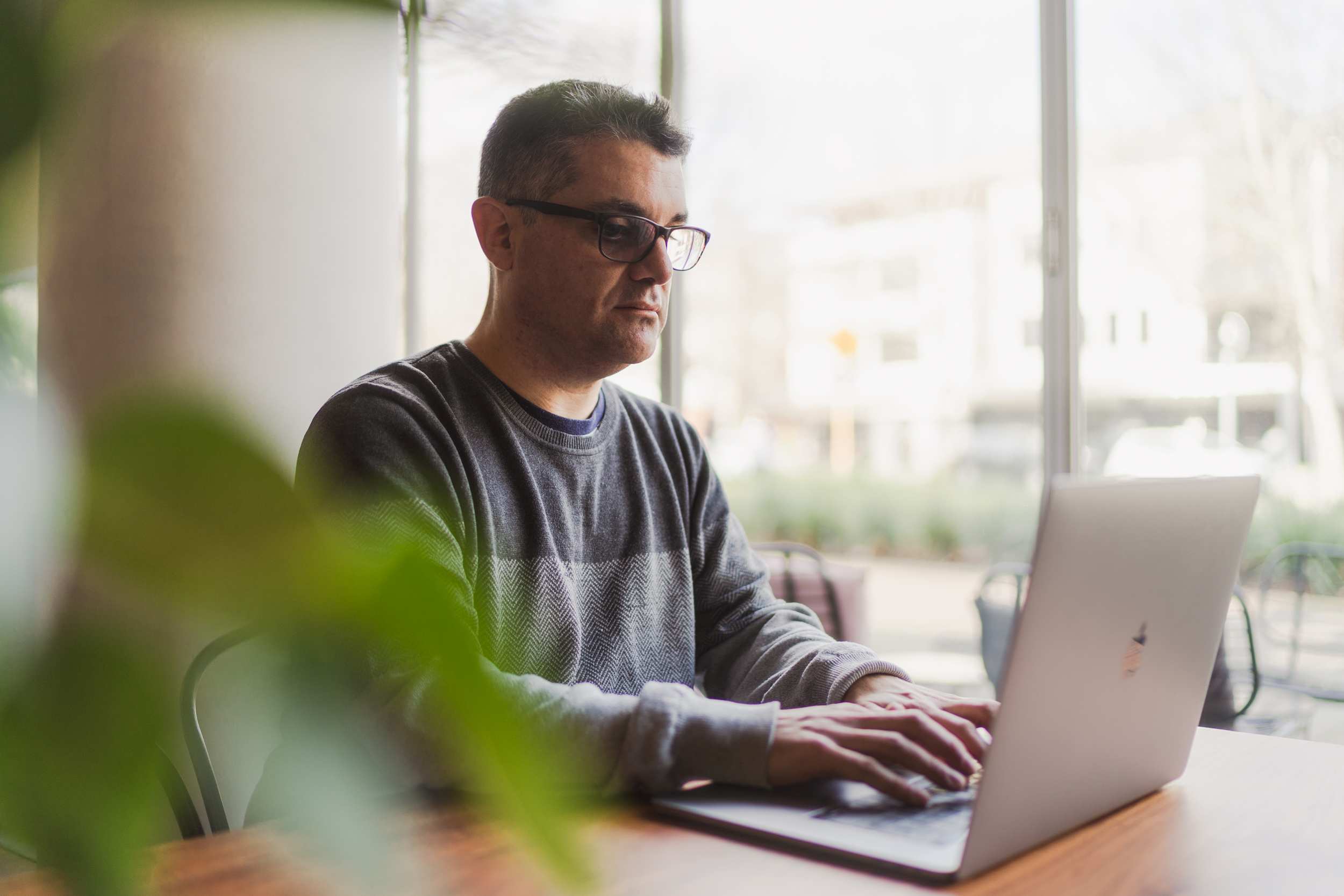 A man with glasses and a grey jumper sits at a desk working on a laptop next to a window.
