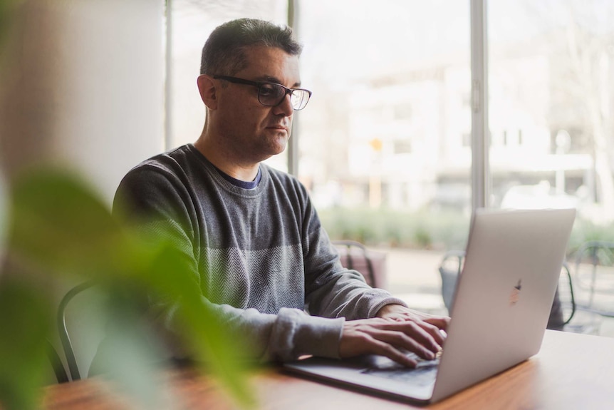 A man with glasses and a gray sweater sits at a desk and works on a laptop next to a window.