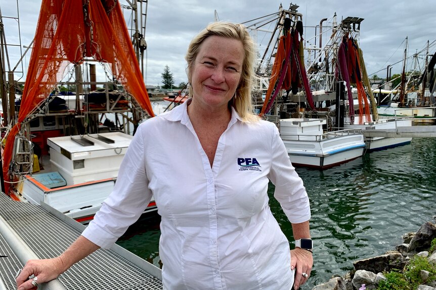 A woman in a white shirt stands in front of a fleet of prawn trawlers.