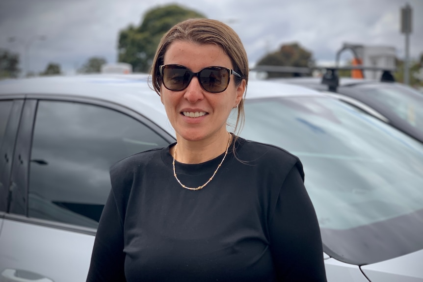 Lena is wearing dark sunglasses and standing next to a car outdoors.