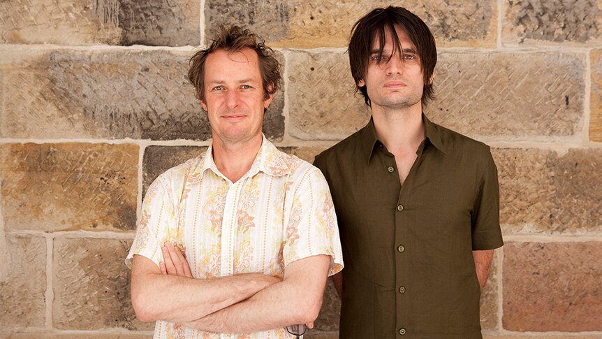 A photograph of Richard Tognetti and Jonny Greenwood side by side in front of a sandstone wall.