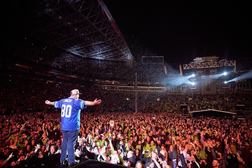 A singer standing on stage in front of a large audience wearing a blue sports jersey, jeans and baseball cap.