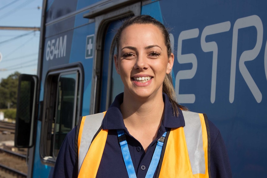 Photo of a young woman smiling and wearing a bright yellow vest and standing in front of a train