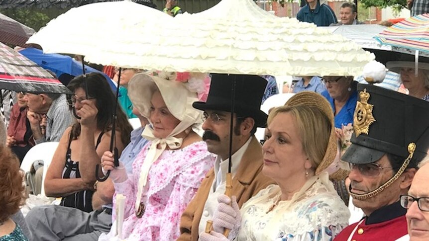 Women earing bonnets and a man in a top hat, under umbrellas.