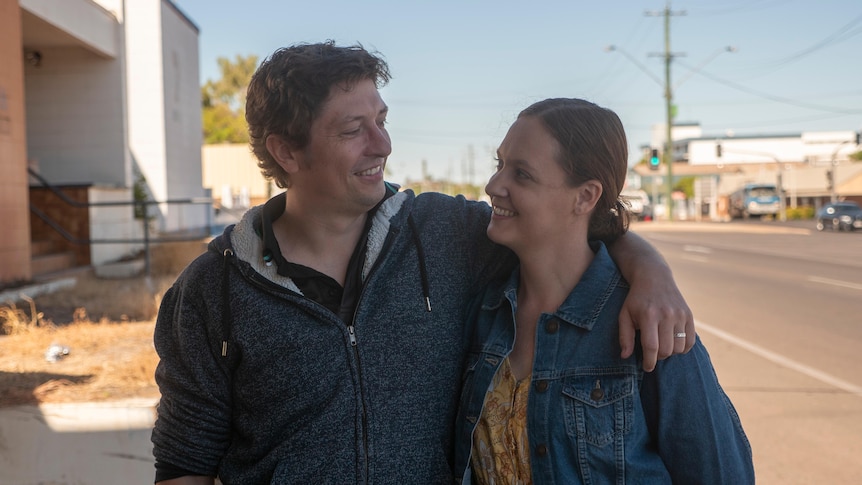 A young couple stand arm in arm outside smiling at each other.