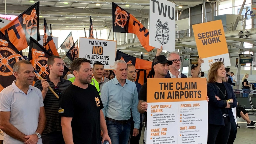 A group of people holding black and orange Transport Workers Union protest signs and flags at an airport.