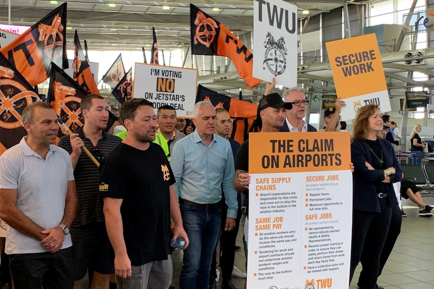A group of people holding black and orange Transport Workers Union protest signs and flags at an airport.