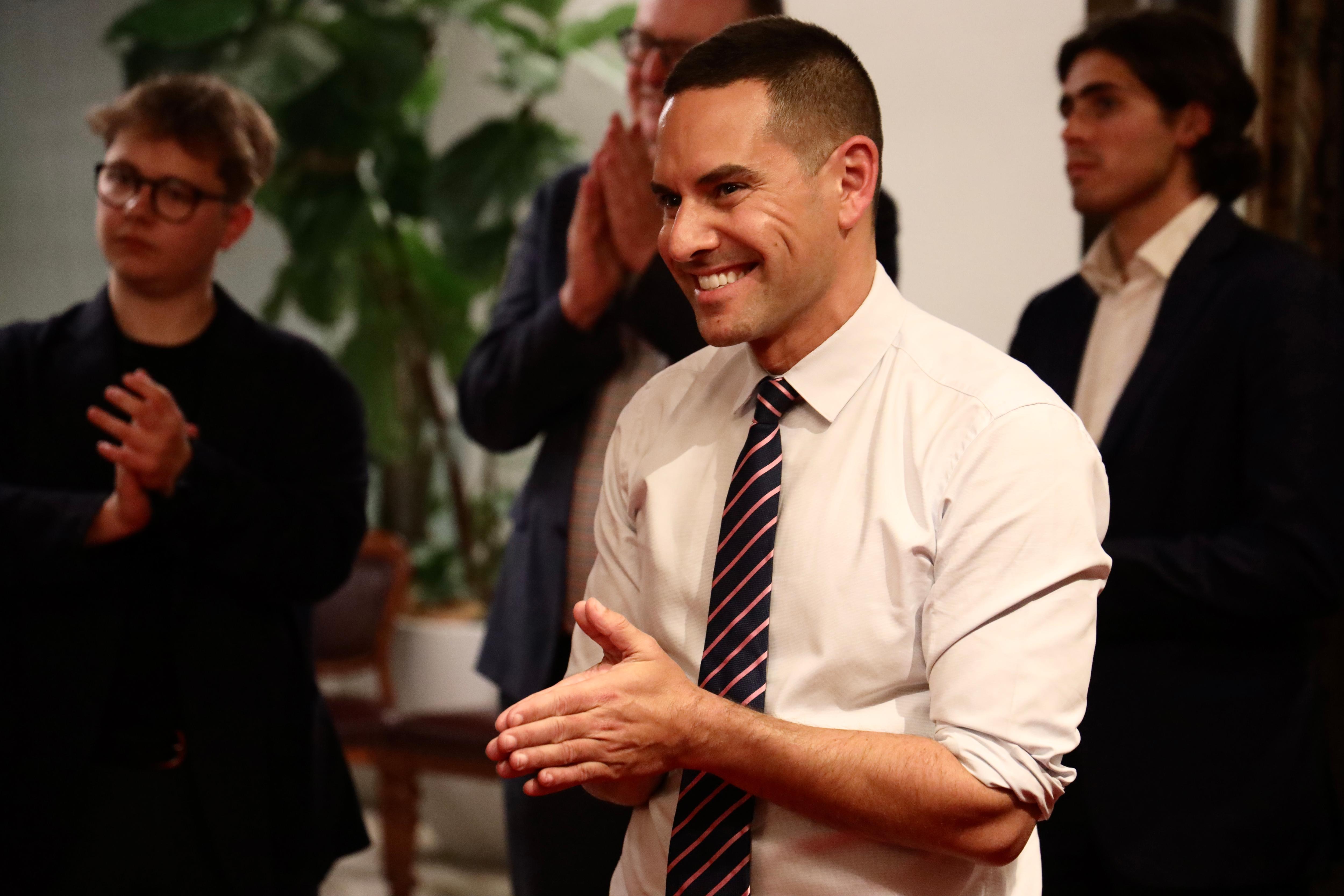 A man in a long sleeve white button-up and a tie smiles.
