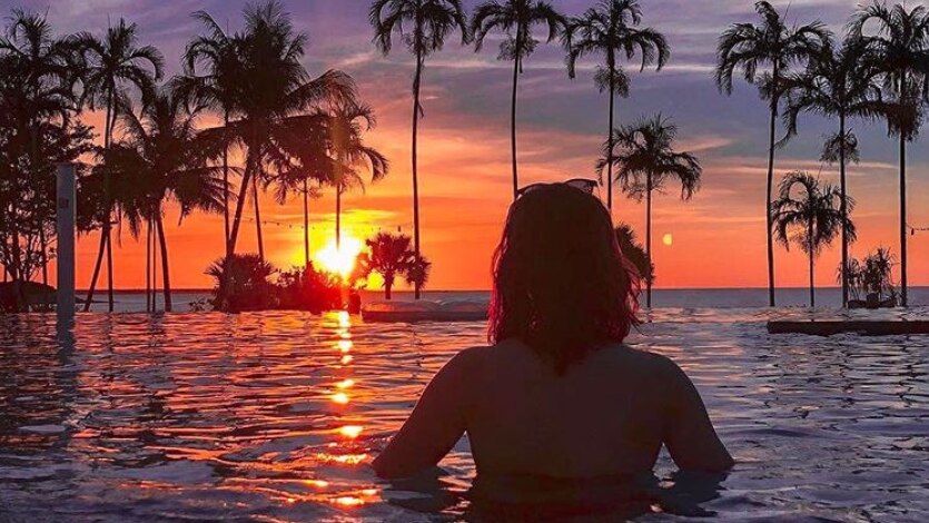 Silhouette of a woman standing chest-deep in water looking at a palm-tree lined sunset from a beachside infinity pool.